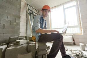 Handsome architect or foreman in helmet working with laptop on the apartment drawings at the structure white interior photo