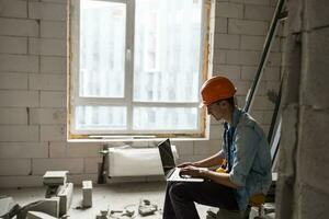 Handsome architect or foreman in helmet working with laptop on the apartment drawings at the structure white interior photo