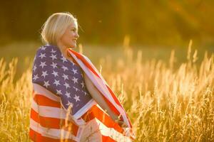 hermosa joven niña participación un americano bandera en el viento en un campo de centeno. verano paisaje en contra el azul cielo. horizontal orientación. foto