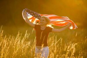 hermosa joven niña participación un americano bandera en el viento en un campo de centeno. verano paisaje en contra el azul cielo. horizontal orientación. foto