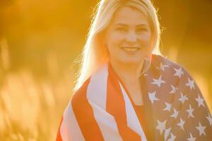 Beautiful young girl holding an American flag in the wind in a field of rye. Summer landscape against the blue sky. Horizontal orientation. photo