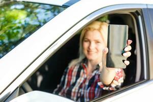 hermosa mujer sentado en un coche y demostración su teléfono inteligente fuera el ventana foto
