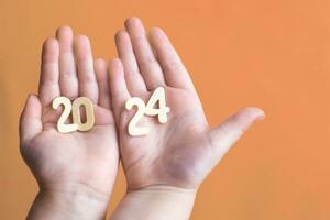 Child hands holding wooden number 2024 on palms on festive Peach Fuzz background with copy space photo