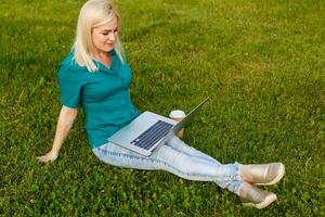 Top view of woman sitting in park on the green grass with laptop, hands on keyboard. Computer screen mockup. Student studying outdoors. Copy space for text photo
