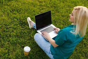 Top view of woman sitting in park on the green grass with laptop, hands on keyboard. Computer screen mockup. Student studying outdoors. Copy space for text photo