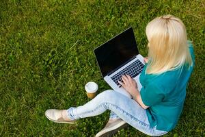 Top view of woman sitting in park on the green grass with laptop, hands on keyboard. Computer screen mockup. Student studying outdoors. Copy space for text photo