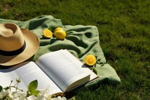 ai generado almuerzo descanso en el parque. picnic cobija con un abierto libro, limones, playa bolso y ancho borde Paja sombrero en un jugoso verde recién recién cortado césped. generativo ai. foto