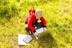 young boy with a books and laptop computer on green grass in the park photo