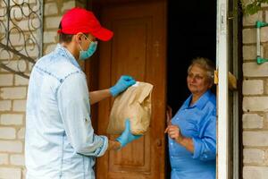 Young male volunteer in mask gives an elderly woman boxes with food near her house. Son man helps a single elderly mother. Family support, caring. Quarantined, isolated. Coronavirus covid-19. Donation photo