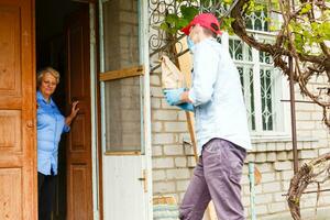 Young male volunteer in mask gives an elderly woman boxes with food near her house. Son man helps a single elderly mother. Family support, caring. Quarantined, isolated. Coronavirus covid-19. Donation photo