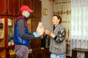 Young male volunteer in mask gives an elderly woman boxes with food near her house. Son man helps a single elderly mother. Family support, caring. Quarantined, isolated. Coronavirus covid-19. Donation photo