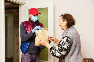 Young male volunteer in mask gives an elderly woman boxes with food near her house. Son man helps a single elderly mother. Family support, caring. Quarantined, isolated. Coronavirus covid-19. Donation photo