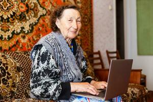 antiguo mujer trabajando en ordenador portátil computadora a hogar, abuela utilizando cuaderno y buscando en Internet sitio foto