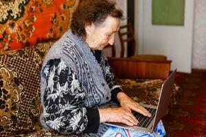 antiguo mujer trabajando en ordenador portátil computadora a hogar, abuela utilizando cuaderno y buscando en Internet sitio foto