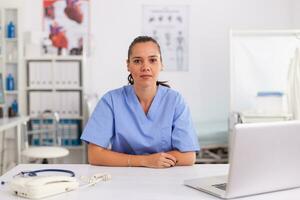 Portrait of pretty medical nurse smiling at camera in hospital office wearing blue uniform. Healthcare practitioner sitting at desk using computer in modern clinic looking at monitor, medicine. photo