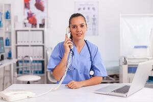 Portrait of medical nurse smiling at camera while using phone in hospital office. Health care physician sitting at desk using computer in modern clinic looking at monitor. photo