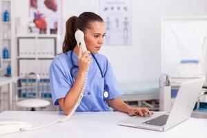 médico facultativo respondiendo teléfono llamadas y Planificación equipo en hospital oficina. salud cuidado médico sentado a escritorio utilizando computadora en moderno clínica mirando a monitor. foto