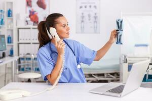 Medical nurse holding patient radiography in hospital office while talking with doctor on phone. Health care physician sitting at desk using computer in modern clinic looking at monitor. photo