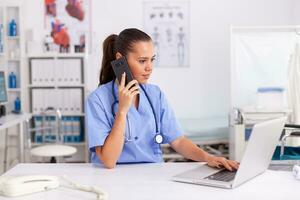 Medical practitioner using telephone and laptop in hospital office wearing blue uniform. Health care physician sitting at desk using computer in modern clinic looking at monitor. photo