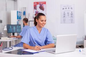 Medical practitioner sitting at desk in hospital office using laptop with doctor in the background. Health care physician using computer in modern clinic looking at monitor, medicine, profession. photo