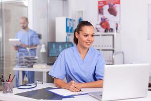 Smiling nurse using laptop computer and writing notes on clipboard in hospital office with doctor in the background. Health care physician using computer in modern clinic looking at monitor, photo