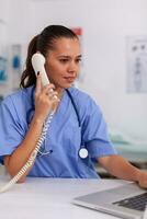 Medical practitioner having a conversation with patient on phone from hospital office and checking appointment. Health care physician sitting at desk using computer in modern clinic looking at monitor. photo