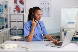 Medical practitioner using telephone and laptop in hospital office wearing blue uniform. Health care physician sitting at desk using computer in modern clinic looking at monitor. photo