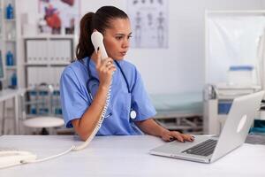 médico facultativo respondiendo teléfono llamadas y Planificación equipo en hospital oficina. salud cuidado médico sentado a escritorio utilizando computadora en moderno clínica mirando a monitor. foto