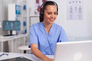 Medical staff talking with patient about prescription wearing headset with microphone in hospital office. Health care physician sitting at desk using computer in modern clinic looking at monitor. photo