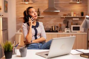 Freelance woman having a phone coversation with a client late at night from home kitchen. Employee using modern technology at midnight doing overtime for job, business, career, network, lifestyle. photo