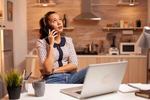 Lanza libre mujer teniendo un teléfono cobertura con un cliente tarde a noche desde hogar cocina. empleado utilizando moderno tecnología a medianoche haciendo tiempo extraordinario para trabajo, negocio, carrera profesional, red, estilo de vida. foto