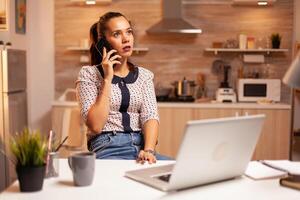 concentrado mujer de negocios durante teléfono llamada tarde a noche desde hogar oficina. empleado utilizando moderno tecnología a medianoche haciendo tiempo extraordinario para trabajo, negocio, ocupado, carrera profesional, red, estilo de vida ,inalámbrico. foto