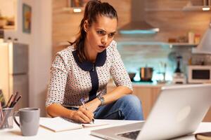 Businesswoman writing notes while working late on computer in home kitchen. Employee using modern technology at midnight doing overtime for job, business, busy, career, network, lifestyle ,wireless. photo