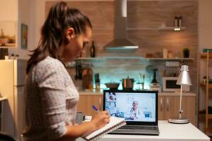 Woman having a video conference with doctor late at night and taking notes. Physician giving consultation to sick patient from hospital office during virtual examination, device, medicine, appointment. photo