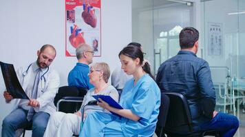 Medical staff discussing diagnosis with disabled senior woman in wheelchair in hospital hallway. Old man waiting for medical examination. Nurse taking notes on clipboard. photo