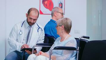 Doctor discussing diagnosis and recovery treatment with disabled senior woman in wheelchair. Old man in hospital waiting area. Medic wearing stethoscope and white coat. photo