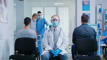 Doctor with visor against coronavirus in waiting area looking intro camera. Nurse with face mask assisting disabled woman with walking frame in hospital hallway. photo