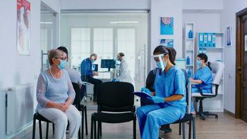 Nurse and senior patient going in hospital examination room from waiting area. Doctor inviting old woman for consultation in office. Wearing mask against coronavirus. photo