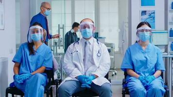 Team of medical staff with protection mask and visor against coronavirus in hospital waiting area looking at camera. Patient in examination room for consultation. photo