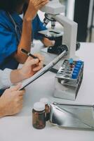 Medical worker in lab coat and sterile mask, doing a microscope analysis while her colleague are working behind photo