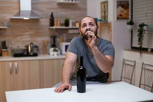 Pensive man drinking glass of wine while sitting alone and depressed. Person with alcohol addiction having bottle of alcoholic beverage, liquor, booze, feeling heartbroken and desperate photo