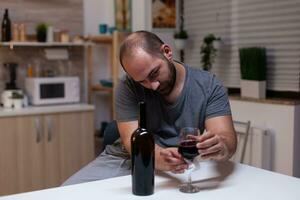 Caucasian man holding glass of wine sitting in kitchen at home. Depressed person drinking liquor, booze, alcoholic beverage alone while feeling intoxicated with bottle of alcohol photo