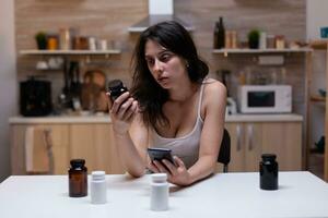 retrato de mujer con doloroso migraña participación botella de medicina leyendo prescripción para pastillas, drogas, recurso, medicamento, tratamiento y medicamento en teléfono inteligente en contra dolor de cabeza foto