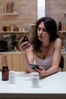 Sick woman with migraine looking at container with medicine and smartphone for prescription. Person with headache holding bottle of drugs, medication, remedy, medicament, treatment photo