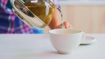 Green tea flowing from teapot in slow motion. Close up of tea from the kettle slowly pour into porcelain cup in the kitchen in the morning at breakfast, using teacup and healty herbal leaves. photo