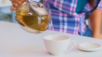 Close up of tea from the kettle slowly pour into porcelain cup. Young lady preparing green tea in the kitchen in the morning at breakfast, using teapot, teacup and healty herbal leaves. photo