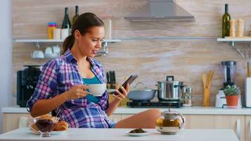 Woman using smart phone for social media and during breakfast drinking a cup of green tea in kitchen. Holding phone device with touchscreen using internet technology scrolling, searching on gadget photo