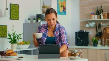 Portrait of young woman using tablet in the morning sitting at the table in the kitchen drinking tea. Working from home using device with internet technology, typing, on gadget during breakfast. photo