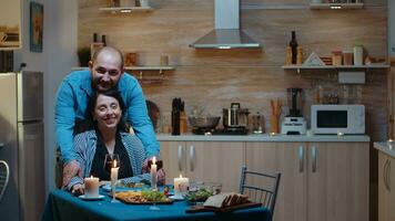 portret de contento joven Pareja sonriente a cámara sentado a el mesa en el cocina durante romántico cena. casado personas especial oferta momentos, disfrutando el comida a vela luces celebracion foto