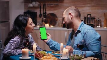 Young happy couple holding green screen phone at dinner. Cheerful people looking at mockup template chroma key isolated smart phone display using techology internet sitting at the table in kitchen. photo